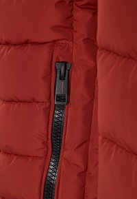 Chaqueta acolchada roja con una textura brillante y acolchada. Cuenta con un cierre negro con una lengüeta rectangular y costuras visibles.