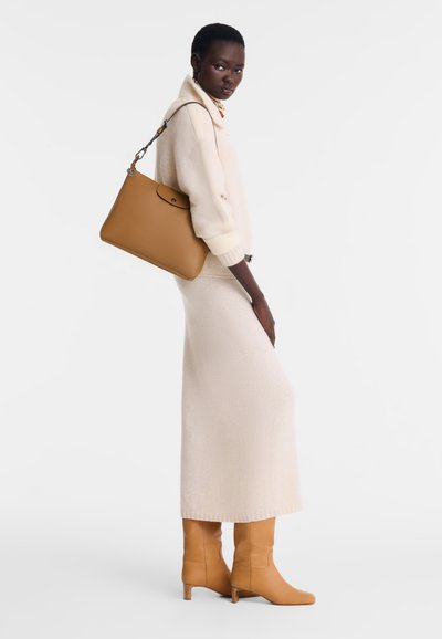 Tan leather handbag with rounded shape and a flap closure. Model in cream sweater and skirt, paired with knee-high tan boots.