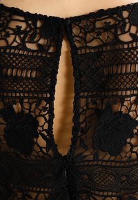 Musta pitsipaita, jossa on syvä V-malli selässä, monimutkaisia kukkakuvioita ja vetoketju. Pehmeä, läpikuultava rakenne yksityiskohtaisella brodeerauksella.