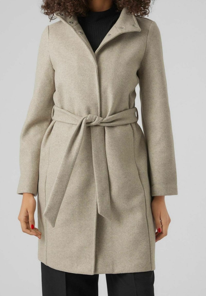 Femme portant un manteau en laine beige ceinturé par-dessus un haut noir et un pantalon noir sur un fond uni gris clair.