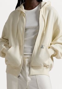 Beżowa jasna bluza z kapturem zapinana na zamek, z bocznymi kieszeniami, ściągaczami na rękawach i kapturem ze sznurkiem. Nosi się ją na biały prążkowany top. Naszywka z logo z przodu.