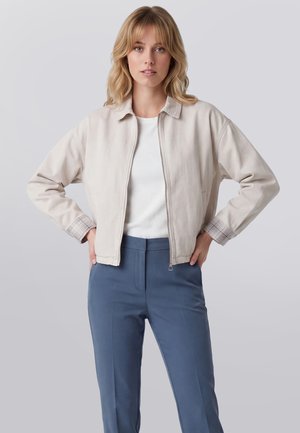 Junge Frau mit blondem Haar, die eine hellbeige Zip-up-Jacke, ein weißes Hemd und blaue Hosen trägt und mit Händen in der Hüfte steht.
