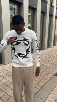 Strickpullover in Off-White mit einem schwarzen Flammenmuster und gerippten Bündchen, kombiniert mit beigen Hosen auf einer mit Backsteinen gepflasterten Fläche.