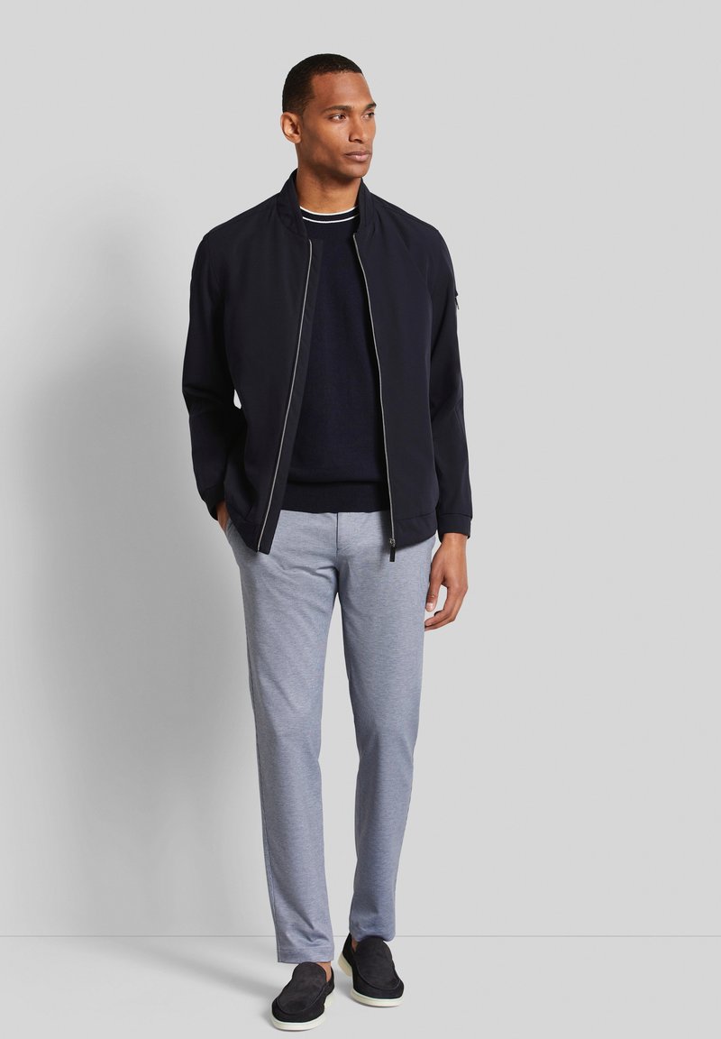 Veste de bombardier navy avec fermeture éclair, pull gris, pantalon gris clair et chaussures slip-on noires, sur fond neutre.