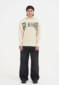 Cremfarbener, gerippter Strickhoodie mit schwarzem Grafiktext, kombiniert mit lockeren schwarzen Denimhosen mit strukturierten Akzenten und weitem Bein.