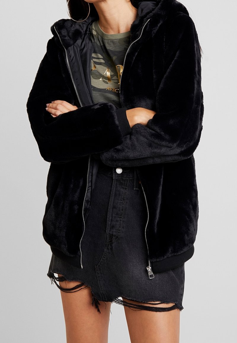 Sort kunstpels hættejakke med lynlås, ribbede manchetter og en tekstureret finish, båret over en grøn camouflage T-shirt og en slidset sort nederdel.