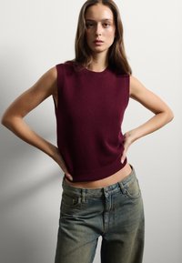Top sin mangas de punto burdeos con cuello y dobladillo acanalados, combinado con jeans de cintura alta en azul claro. Fondo neutro.