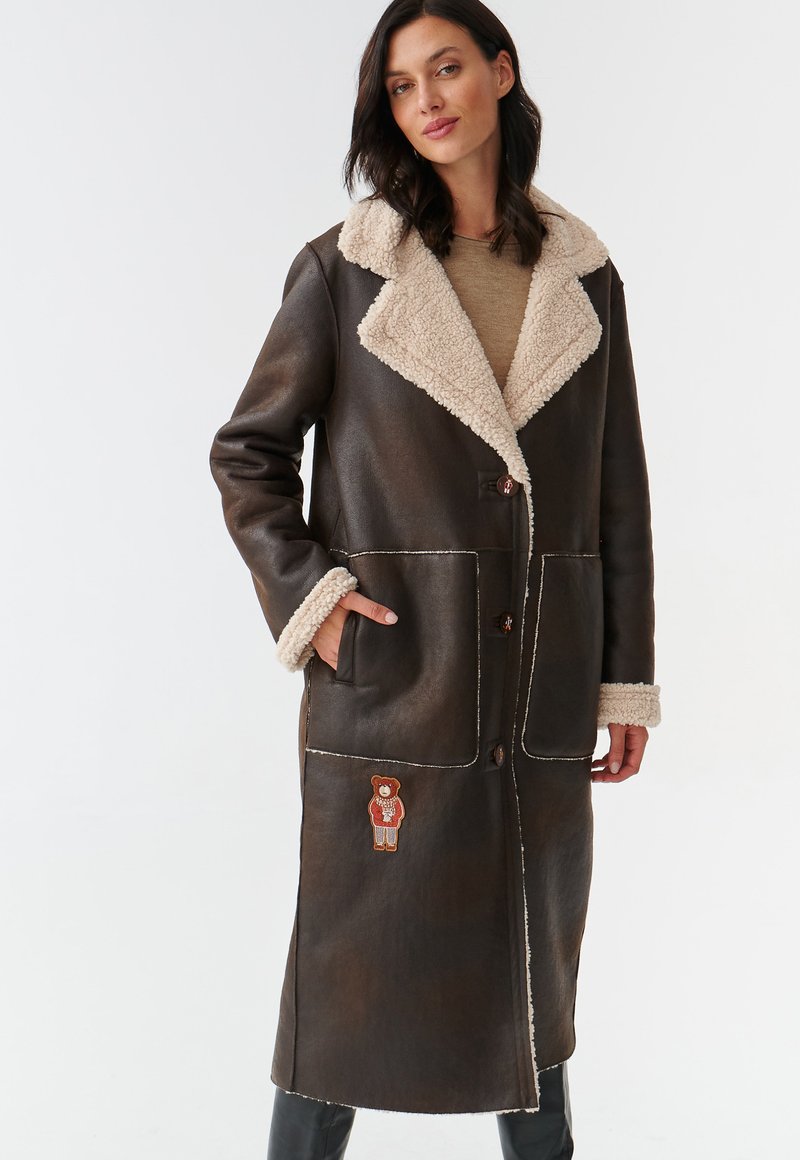 Manteau en cuir marron avec un col en fausse fourrure beige, grandes poches, fermeture à boutons et un motif de ours brodé sur le bas à l'avant.