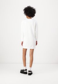 Abito felpa bianco con maniche lunghe, scollatura arrotondata e spacco laterale; abbinato a scarpe con piattaforma nere e calzini bianchi.