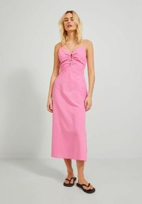 Vestido midi rosa con un corpiño ajustado, detalle fruncido en el centro y tirantes finos. Combinado con chanclas negras sobre un fondo simple.