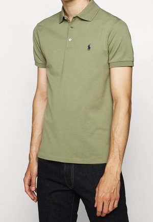 Olivengroene polo met een klassieke kraag, korte mouwen, een drieknopenrij en een marineblauw geborduurd logo op de borst. Draagt het met een donkere spijkerbroek.