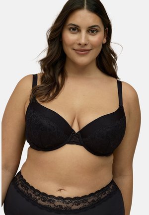 Lachende vrouw met golvend bruin haar, gekleed in een zwarte kanten beha en bijpassend hooggetailleerd ondergoed, tegen een witte achtergrond.