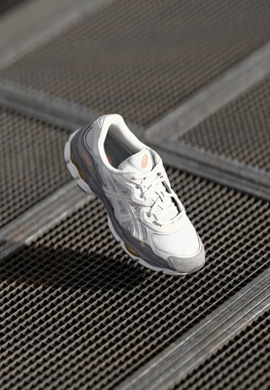 Weißer und grauer Asics Laufschuh schwebt über einer Metallgitterfläche mit diagonalen Linien im weichen Tageslicht.