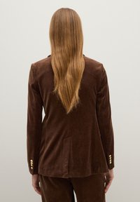 Bruine fluwelen blazer met een getailleerde pasvorm, voorzien van een enkele split, vier gouden knopen op de mouwen en een gladde textuur.