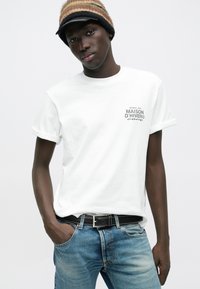 Νεαρός άνδρας που φοράει λευκό μπλουζάκι Maison d'Hiver, μπλε τζιν, μαύρη ζώνη και ριγέ σκουφάκι, στέκεται με το ένα χέρι στην τσέπη.