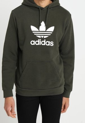 Persoon draagt een donkergroene Adidas hoodie met wit logo en kangoeroezak, gecombineerd met een zwarte broek.