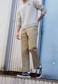 Šedá mikina s žebrovanými manžetami, béžové chinos kalhoty a černé tenisky Vans. Oblečení má jednoduchý design a volný střih, bez viditelných vzorů.