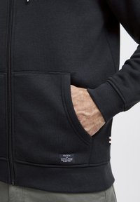 Haut à capuche zippé noir en tissu doux, avec des poches kangourou et des poignets côtelés. Étiquette de marque discrète cousue à l'ourlet, design minimaliste.