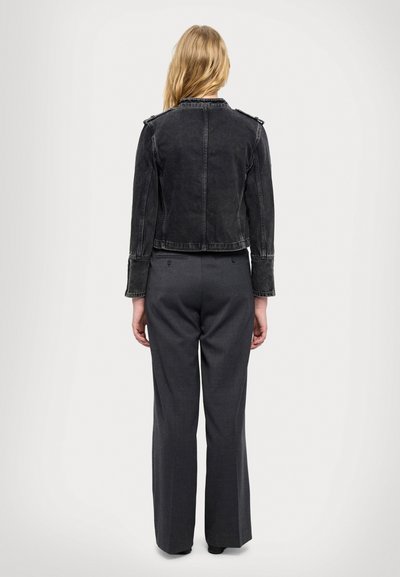 Šviesiaplaukė moteris stovi atsukusi nugarą, vilki trumpą juodą džinsinę striukę ir tamsiai pilkus, aukštu liemeniu ir plačiomis kelnėmis.