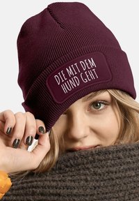 Burgundy knit beanie with a textured finish, featuring a rectangular patch with white text reading “DIE MIT DEM HUND GEHT.”