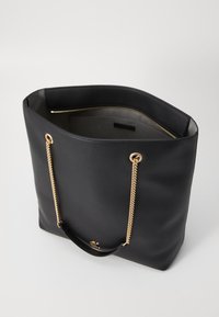 Black leather tote bag with gold metal chain accents, a smooth texture, and open top. Interior is lined and features an interior pocket.