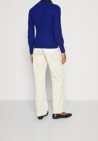 Pull bleu en maille à manches longues, texture côtelée, et taille ajustée, associé à un pantalon large crème et des chaussures plates noires.