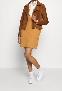 Braune Wildleder-Bikerjacke über einem senfgelben Kleid mit Fronttaschen, kombiniert mit weißen Sneakers und Knöchelsocken.