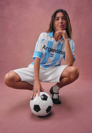 Žena v argentínskom futbalovom drese drepuje s jednou rukou na futbalovej lopte na jednoliatom ružovom pozadí.