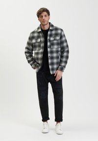 Giacca a motivo check bianco e nero, design con bottoni, due tasche frontali, indossata su un maglione nero, abbinata a pantaloni neri strappati e sneakers bianche.