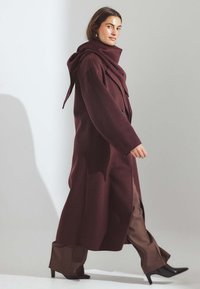 Cappotto bordeaux con cappuccio, design doppiopetto e tasche laterali, abbinato a pantaloni a gamba larga marroni e scarpe nere con tacco.