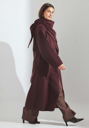 Cappotto bordeaux con cappuccio, design doppiopetto e tasche laterali, abbinato a pantaloni a gamba larga marroni e scarpe nere con tacco.