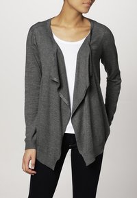 Kobieta ubrana w ciemnoszary kardigan zapinany z przodu, pod który ma na sobie białą bluzkę z okrągłym dekoltem i czarne spodnie, stoi z jedną ręką w luźnej pozycji.