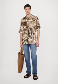 Majica s kratkimi rokavi, bež in črno z zebrastim vzorcem, z rumenim logotipom, v kombinaciji z modrimi kavbojkami in rjavim papirnatim nahrbtnikom.