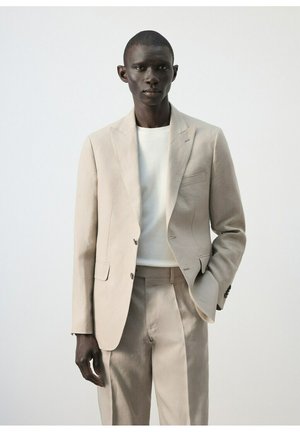 Uomo con un abito beige e una camicia bianca, in piedi con una mano in tasca davanti a uno sfondo chiaro e uniforme.