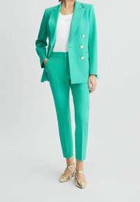 Turkusowa marynarka i dopasowane spodnie z gładkiego materiału, z podwójnymi guzikami i kieszeniami; zestawione ze złotymi sandałami na paski.