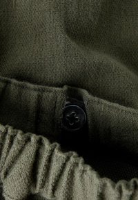 Un tejido verde texturizado presenta un dobladillo fruncido con un acento de botón negro y detalles elásticos, destacando su material suave de punto.