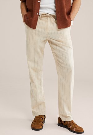 Mand iført beige bukser med snøre og kalkunstriber, brune sandaler med åben tå, hvid t-shirt og rustfarvet kortærmet skjorte med hænderne i lommerne.