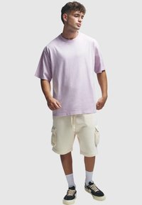 Lilla oversized t-skjorte, myk stoff, rund hals. Kremfargede cargo shorts med sidelommer, avslappet passform. Svarte og kremfargede sneakers, hvite sokker.