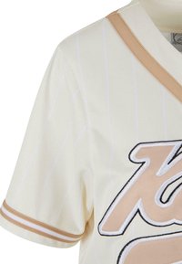 Maglia crema con righe verticali bianche, caratterizzata da un colletto e dettagli beige. Grande logo ricamato in beige e nero sul davanti.