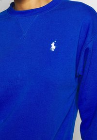 Blå crewneck-tröja gjord av mjukt tyg. Har en liten vit logotyp på bröstet och förstärkt sömnad längs sömmarna.