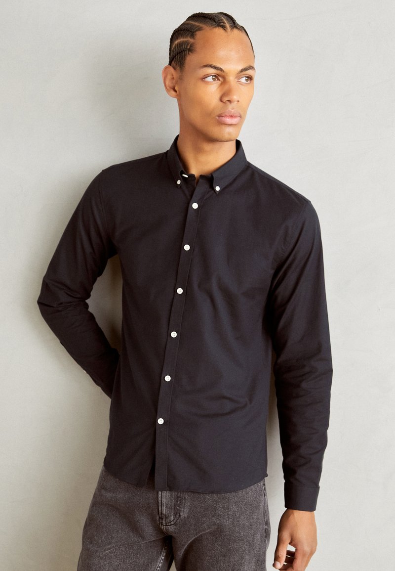 Camisa negra de botones con cuello estructurado, corte slim, mangas largas y botones blancos en contraste. Llevada con pantalones gris oscuro.
