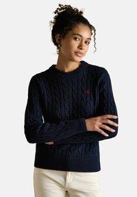 Žena s kudrnatými vlasy, oblečená v námořnickém modrém svetr s copánkovým vzorem a malým červeným logem, v krémových kalhotách, s překříženýma rukama.