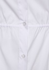 Weißes Hemd mit durchgehender Knopfleiste, gerafftem Oberteil in Taillenhöhe und glattem Stoff. Klare Knöpfe sind sichtbar.