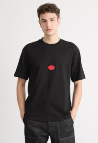 Black crewneck t-shirt made of cotton with short sleeves. Features a red rubber logo patch at the center. Worn with dark denim pants.