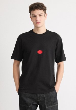 Svart crewneck t-skjorte laget av bomull med korte ermer. Har en rød gummilogg-patch i midten. Brukt med mørke denimbukser.