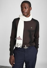 Suéter preto de malha com longa echarpe branca com franjas e "Vivienne Westwood" bordado em bordô. Calças azul marinho com riscas.