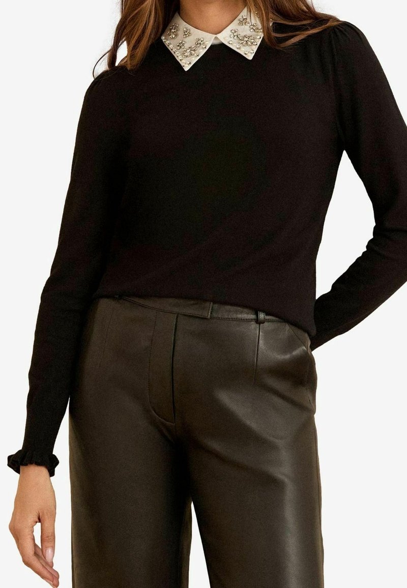 Femme portant un top noir à manches longues avec un col blanc orné et un pantalon en cuir marron foncé taille haute, une main sur la hanche.