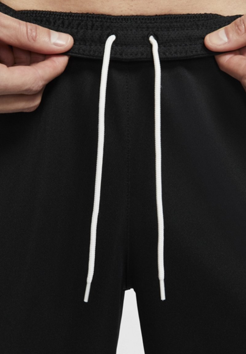 Mains ajustant la ceinture d’un pantalon noir avec de longs lacets blancs centrés à l’avant.