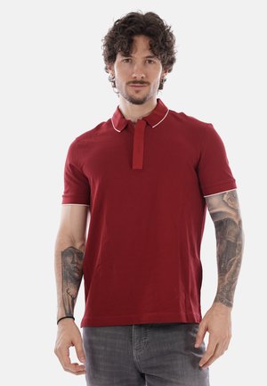 Man with curly hair and tattoos on both arms wearing a red polo shirt and gray jeans, standing against a plain background.