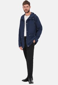 Marineblaue Windjacke mit Kapuze, vorderer Druckknopfleiste, zwei Vorderentaschen und verstellbarem Kordelzug, kombiniert mit schwarzer Hose und Stiefeln.